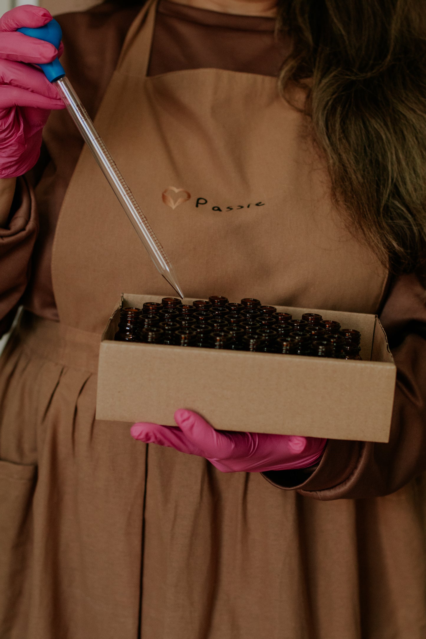 Person wearing a brown apron with 'Passie' branding, holding a box of black objects and a tool.